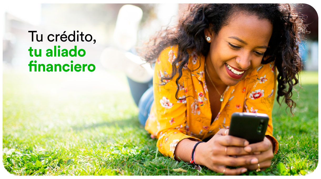 Mujer sonriendo mientras usa su celular recostada en el pasto, con el texto: Tu crédito, tu aliado financiero.