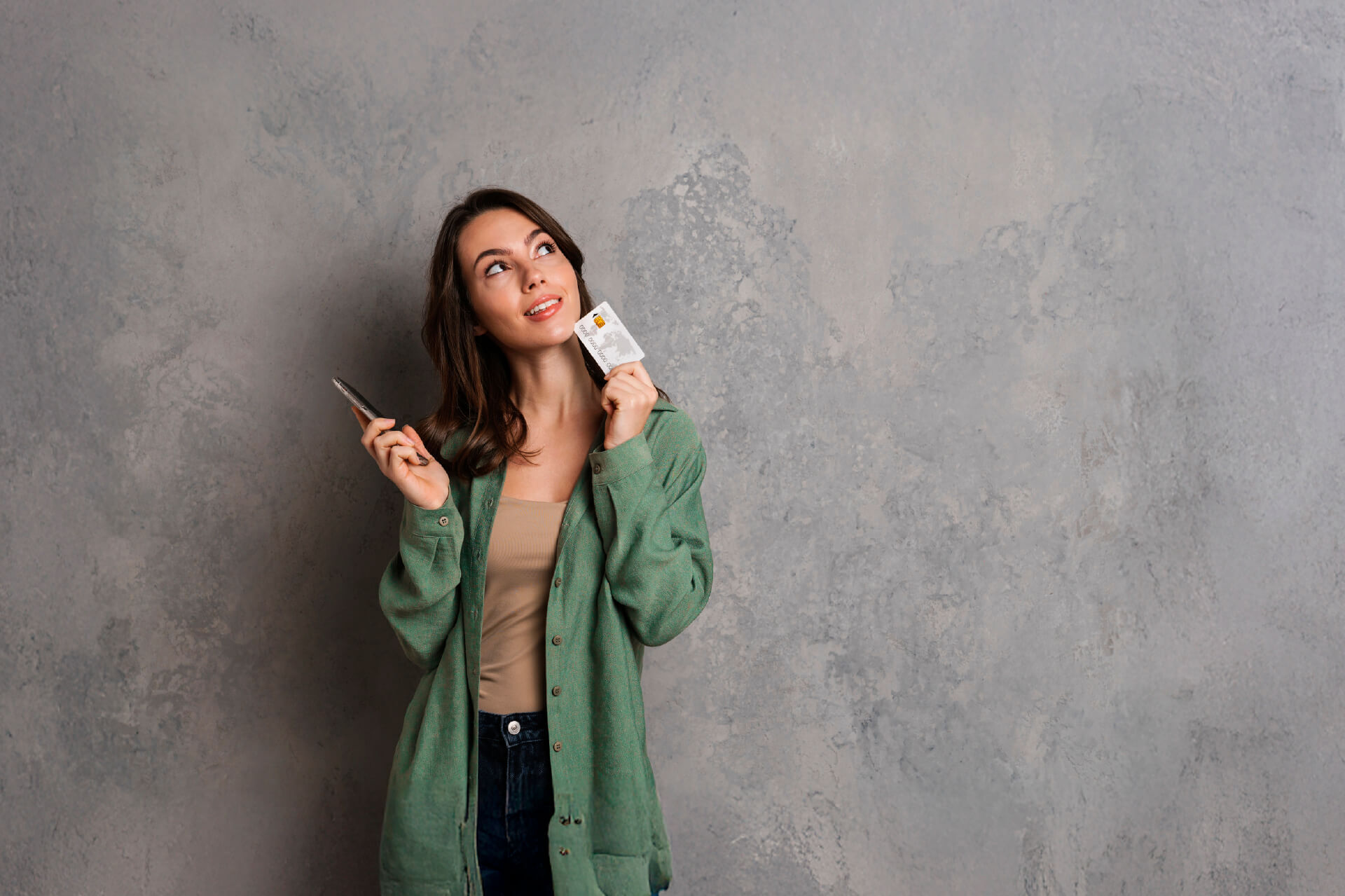 Mujer joven sonriente con camisa verde sostiene un celular en una mano y una tarjeta en la otra, mirando hacia arriba con gesto pensativo frente a una pared gris.