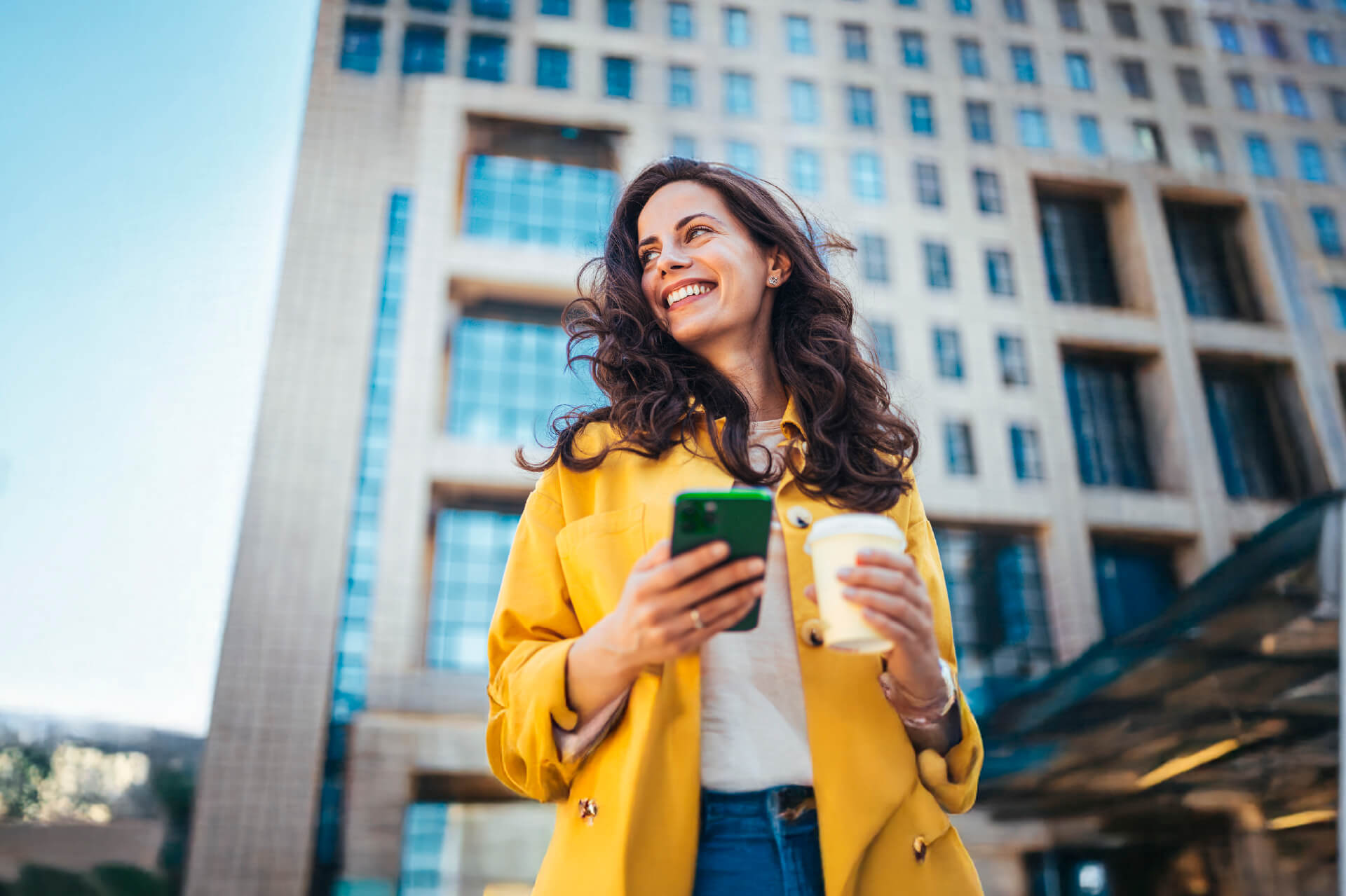Mujer sonriente con abrigo amarillo usando su celular y sosteniendo un café, frente a un edificio corporativo, representando confianza y libertad financiera