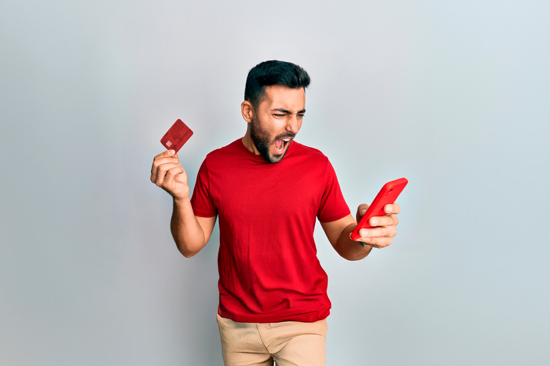 Hombre con camiseta roja sostiene una tarjeta de crédito en una mano y un celular en la otra, mirando la pantalla con expresión de sorpresa o conmoción.