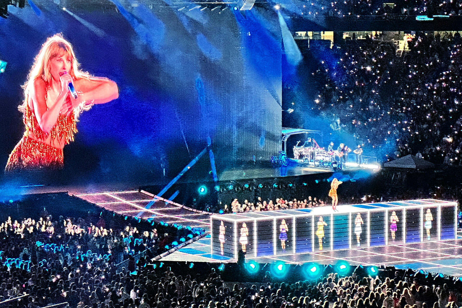Taylor Swift interpreta una canción en el escenario durante su gira, con luces azules, pantalla gigante y miles de fans en el estadio.